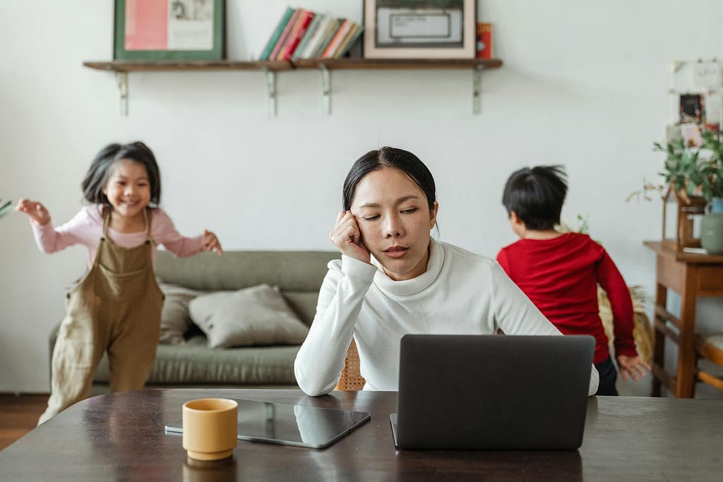 Seorang wanita yang terdistraksi oleh anak-anak di sekitarnya meskipun sedang bekerja di depan laptop.